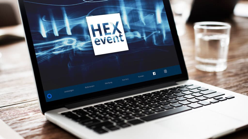 Ein Laptop auf einem Holzschreibtisch zeigt einen Bildschirm mit dem Text „HEX-Ereignis“ und einem blauen abstrakten Muster im Hintergrund. Im Hintergrund steht ein klares Glas Wasser auf dem Schreibtisch, mit verschwommenen Objekten, die eine Büroumgebung suggerieren.