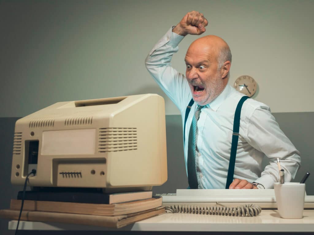 Ein älterer Mann in Oberhemd und Hosenträgern sitzt an einem Schreibtisch und wirkt äußerst frustriert über einen alten Computer. Er hat einen Arm zur Faust erhoben und schreit den Bildschirm an. Auf dem Schreibtisch stehen eine Kaffeetasse und Bücher. An der Wand hinter ihm hängt eine Uhr.