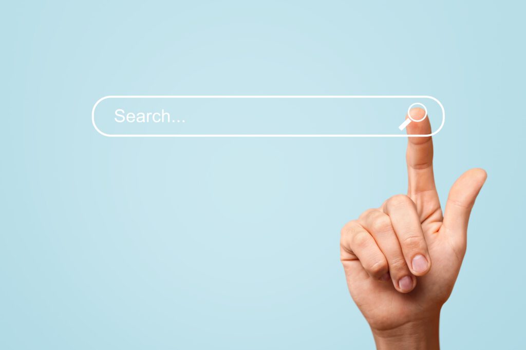 Eine Nahaufnahme einer Hand, die auf eine digitale Suchleiste auf hellblauem Hintergrund zeigt. Die Suchleiste enthält das Wort „Suchen...“ und zeigt ganz rechts, wo der Finger hinzeigt, ein Lupensymbol.