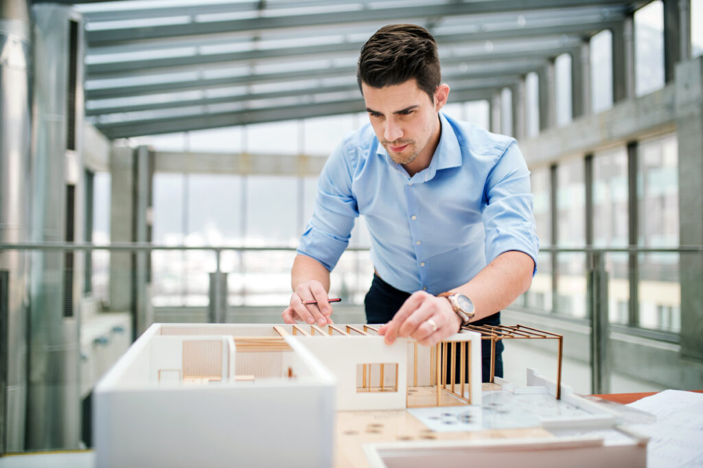 Ein Mann in einem hellblauen Hemd arbeitet in einem geräumigen, modernen Büro mit großen Fenstern sorgfältig an einem detaillierten Architekturmodell. Sein konzentrierter Gesichtsausdruck spiegelt Konzentration wider, während er Teile der Miniaturstruktur anpasst.